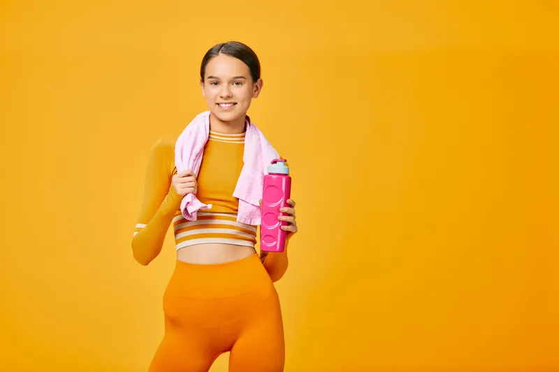 Woman in bright orange athletic wear standing with a towel over her shoulders and a pink water bottle, posed against a vivid orange background, ready for a workout and fitness routine.