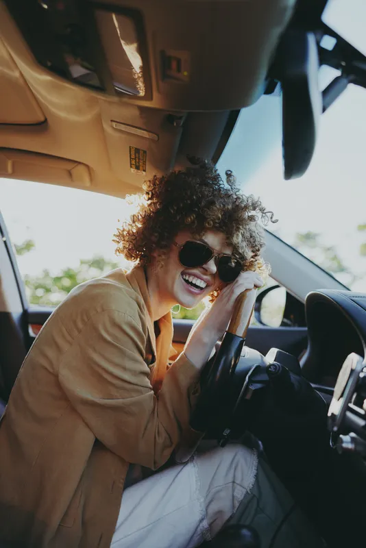 Young woman with curly hair laughs in a sunlit car, wearing a beige top and sunglasses, capturing a carefree road trip moment inside the vehicle, radiating warmth