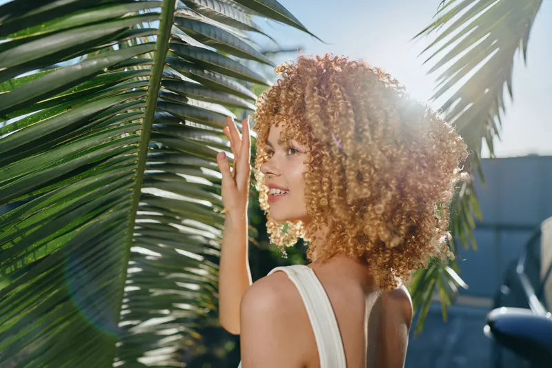 Smiling woman with curly hair touches large green palm leaves outdoors under bright sunlight, showcasing natural beauty and summer vibes in casual sleeveless outfit.