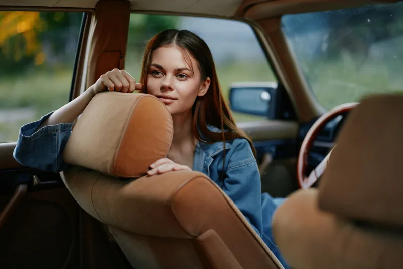 Young woman with brown hair in denim jacket, relaxed inside vintage car, evoking nostalgia and tranquility, captured in soft natural light.