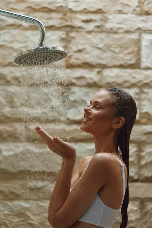 woman enjoying outdoor shower with natural stone wall, wet long hair, relaxed expression, and sunlight highlights, refreshing water flow in warm atmosphere