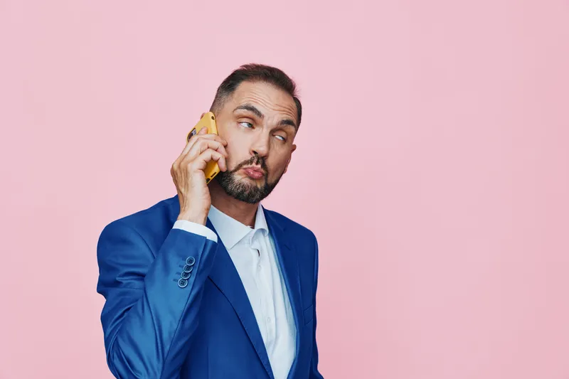 A professional man in a blue suit holds a phone to his ear, standing against a solid pink background. He conveys focus and curiosity, perfect for business scenes that highlight communication and