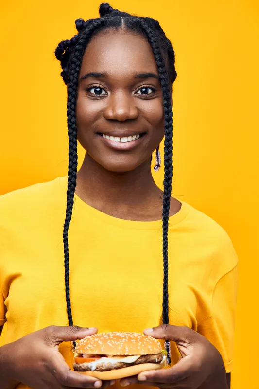 Happy young woman of African descent holding a burger against a bright yellow background, wearing a casual yellow shirt Emphasizes healthy eating and joy