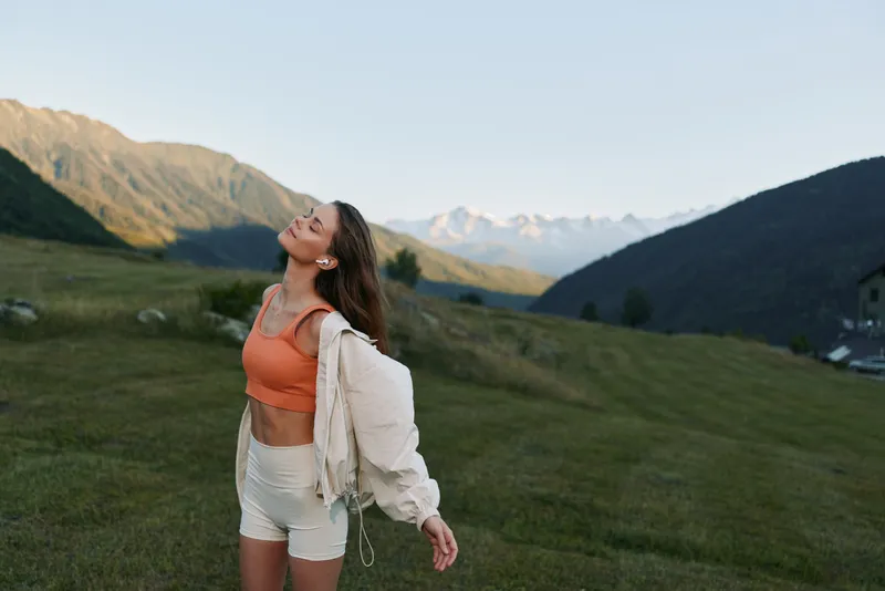 A joyful young woman stands outdoors in a green meadow with distant mountains. She wears an orange crop top and light jacket, enjoying sunlit air and tranquil scenery.