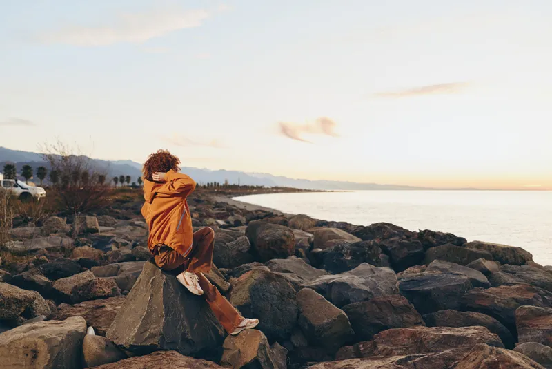 Woman lifestyle outdoors by rocky seashore at sunset, warm evening orange coat and scarf, standing on stones with relaxed posture, soft sea breeze, calm mood, natural colors