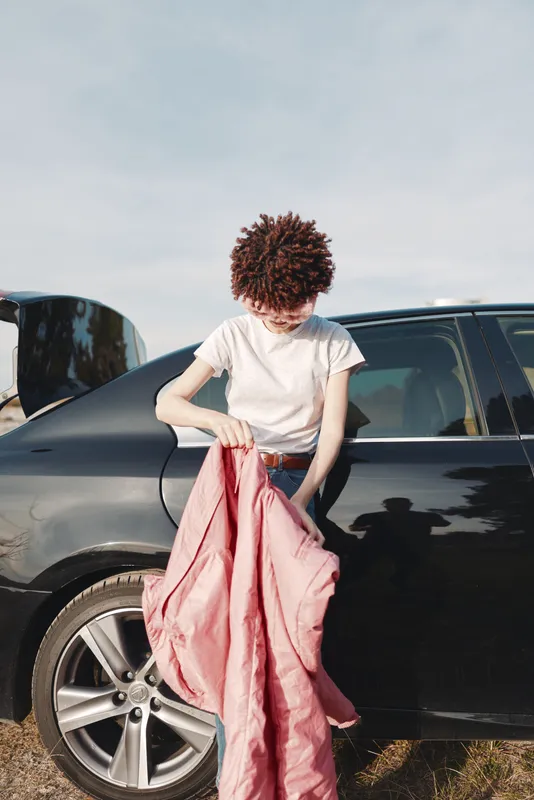 Person with curly hair in a white t-shirt stands beside a dark car, holding a pink garment, outdoor setting, casual fashion moment focusing on clothing and preparation