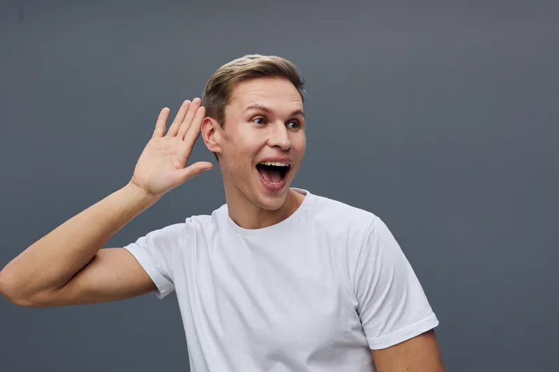 Happy young man with short brown hair wearing white t-shirt posing with hand near ear against gray background. Expressing surprise and joy in studio portrait. People emotion concept, positive mood.
