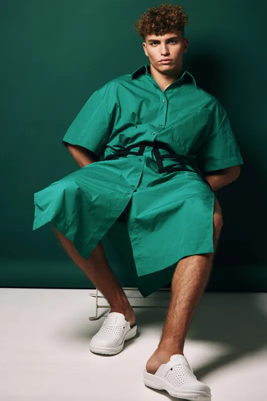 Young man sitting on chair wearing green oversized shirt dress with black belt and white slip-on shoes against dark green background in studio. Fashion, casual style and confident pose.