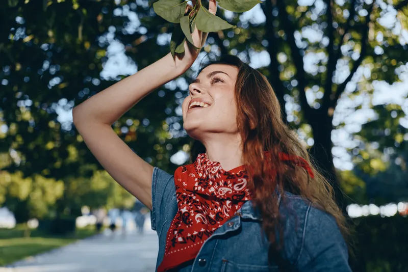 Lifestyle woman in modern boho-western denim street style smiling outdoors with film color effect under green trees on sunny day, casual and vibrant mood.