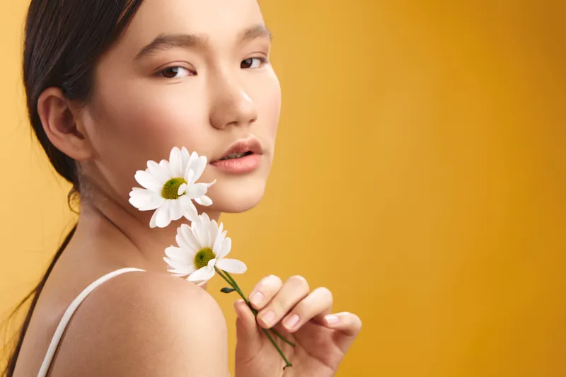 Asian woman gazes softly at the camera, holding white daisies against a warm orange backdrop, creating a natural, fresh portrait that conveys calm confidence and gentle elegance.
