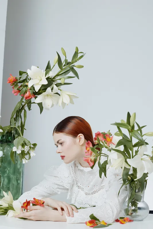 A serene young woman in a white lace blouse surrounded by vibrant flowers, embodying elegance and tranquility, ideal for concepts of beauty and nature