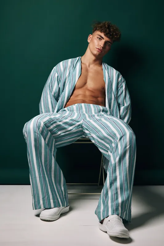 Young man with curly hair wearing striped loose pants and open shirt sitting on chair against dark green background in studio. Casual fashion, relaxed pose and confident expression.