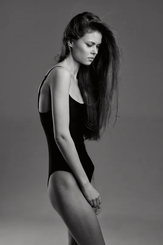 Black and white portrait of a slender woman in a leotard with long hair, expressing pensive mood in minimalistic studio setting.