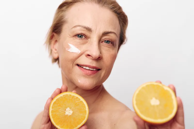 Mature woman with natural skin holds two orange halves near face with cream on cheek. Concept of skincare, healthy lifestyle, and natural beauty with fresh citrus fruit.