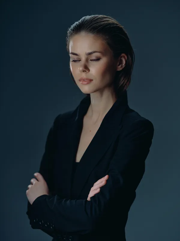 Young woman in black suit with crossed arms and closed eyes showing calm and confidence in dark studio setting