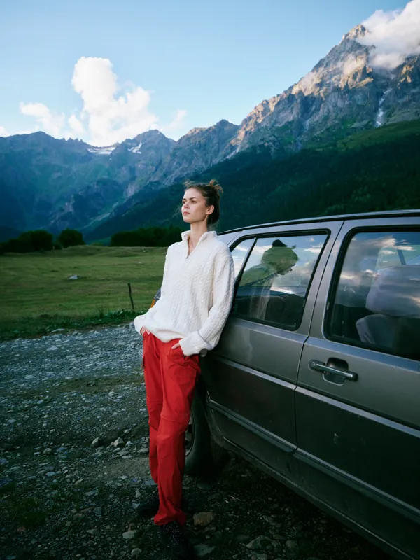 Woman leaning against a car in a mountain landscape, casual outfit with a white blouse and red pants, expansive valley and serene outdoor scene, travel mood