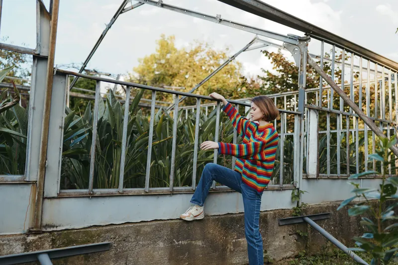Woman in her mid twenties wearing a colorful striped sweater and jeans climbing a greenhouse frame, smiling and curious in an urban garden with tall plants and warm autumn light.