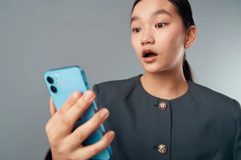 Young woman in dark jacket looking surprised on blue smartphone with open mouth and focused eyes isolated on gray background, communication, technology use and emotional reaction.