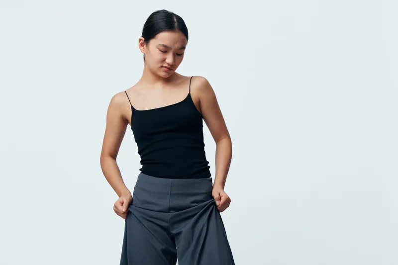 Young woman in black tank top and gray wide pants looking down posing isolated on white background. Casual fashion, modeling, confident pose, studio photo with simple style and natural expression.