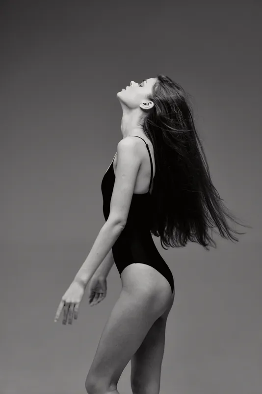 graceful woman in black swimsuit posing with flowing hair against plain background