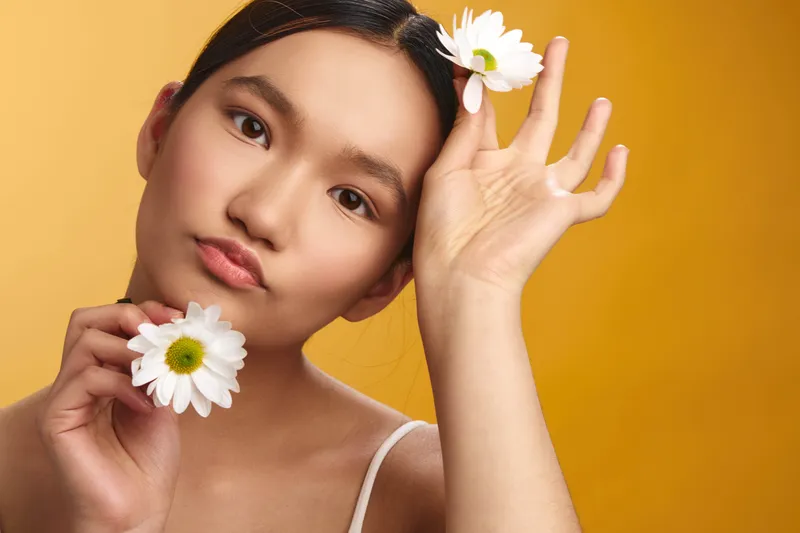 asian woman poses with daisies against a bright yellow backdrop, soft lighting highlights delicate features and a serene expression, creating a fresh floral portrait.