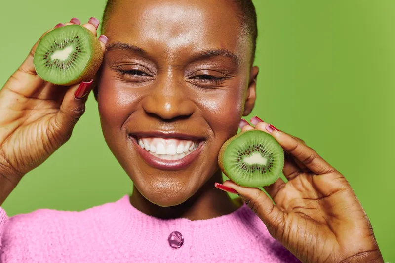 Happy African woman with dark skin holds two halves of fresh kiwi fruit near face. Bright green background and pink sweater create vibrant contrast. Concept of healthy lifestyle and fruit.