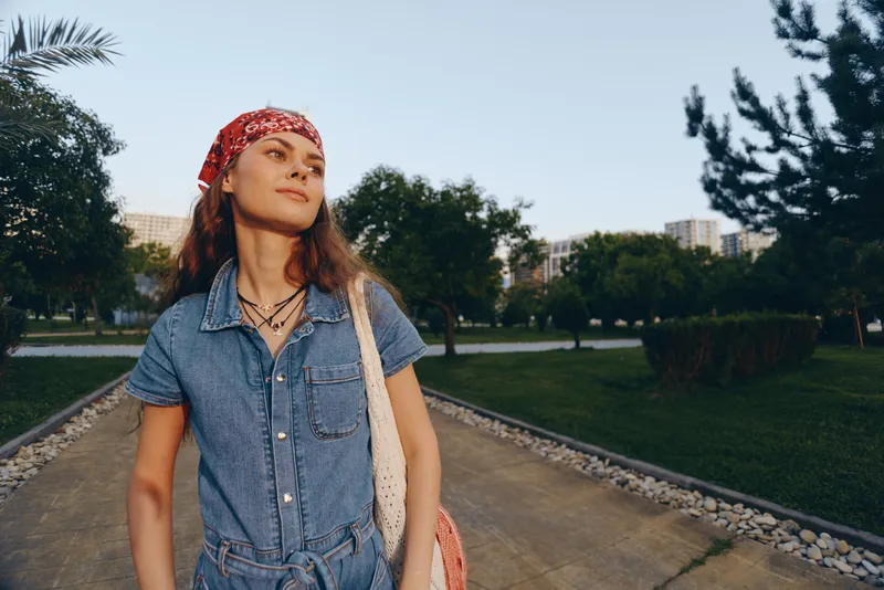young woman wearing denim jumpsuit and red headband walking outdoors in park with green trees and city buildings in background on sunny day