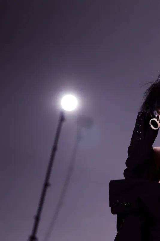 Woman in dark clothing posing near bright streetlight at night with purple background, mysterious atmosphere