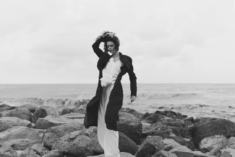 Woman on a rocky coast in a flowing coat, wind-swept hair and a white dress, striking pose by the sea, monochrome fashion image conveying strength, movement and seaside mood