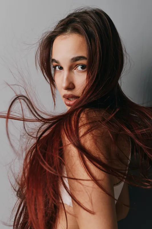 Young woman with long red hair looking over shoulder with serious expression, natural makeup, and soft lighting on neutral background. Fashion portrait with flowing hair movement.