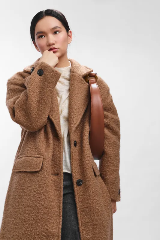 asian woman wearing a textured brown coat and cream knit sweater stands with a thoughtful pose against a neutral backdrop, presenting a stylish urban fashion moment.