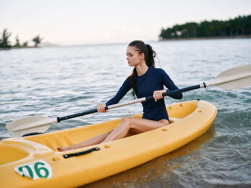 Young woman paddling yellow kayak on calm water with distant trees in soft natural light, enjoying outdoor summer activity and peaceful scenery.