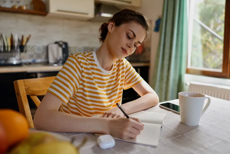 Woman in striped shirt writing in a notebook at a kitchen table, daylight light, mug nearby, cozy home setting, focus on creativity, planning and daily routine.