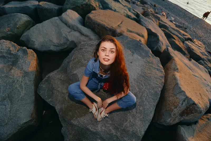 young woman sitting on rocks near water wearing denim overalls with long red hair and relaxed expression at sunset outdoors nature scene