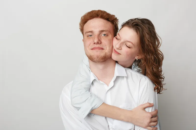 Young couple in white shirts embraces tenderly against a light background. Woman hugs man from behind with closed eyes. Concept of love, affection, intimacy, and relationship.