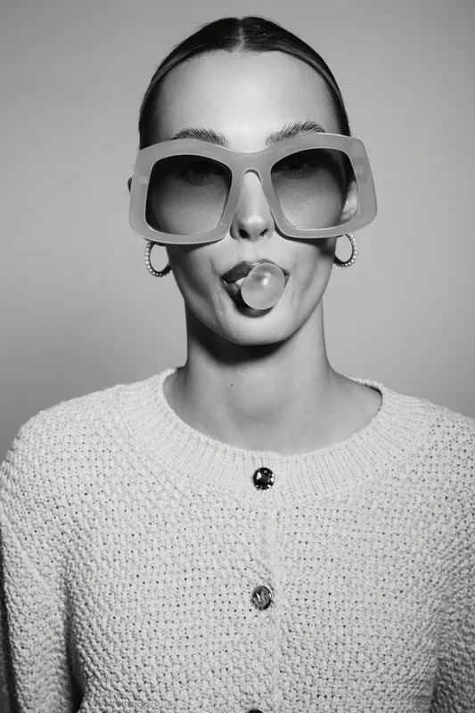 Young white woman with pale skin and slicked hair blows bubblegum bubble wearing oversized sunglasses and knitted sweater in studio.