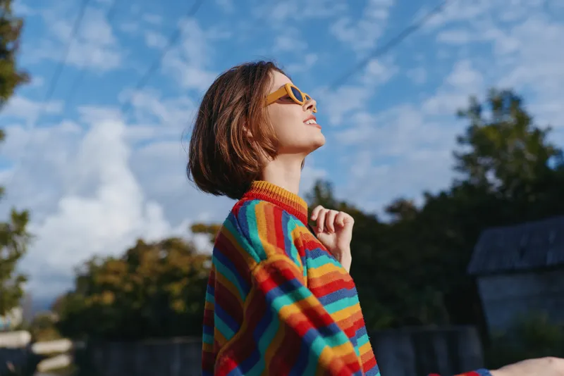 Woman in her twenties wearing a colorful striped sweater and yellow sunglasses smiling outdoors under blue sky, short bob haircut and casual outfit, joyful travel lifestyle portrait.