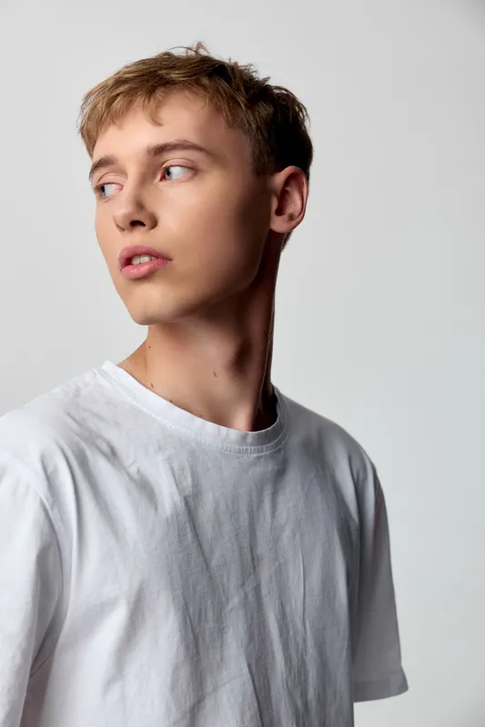 Young man wearing a white shirt in a studio setting, soft lighting, casual upper body pose, looking away for a candid, natural and minimal fashion mood.