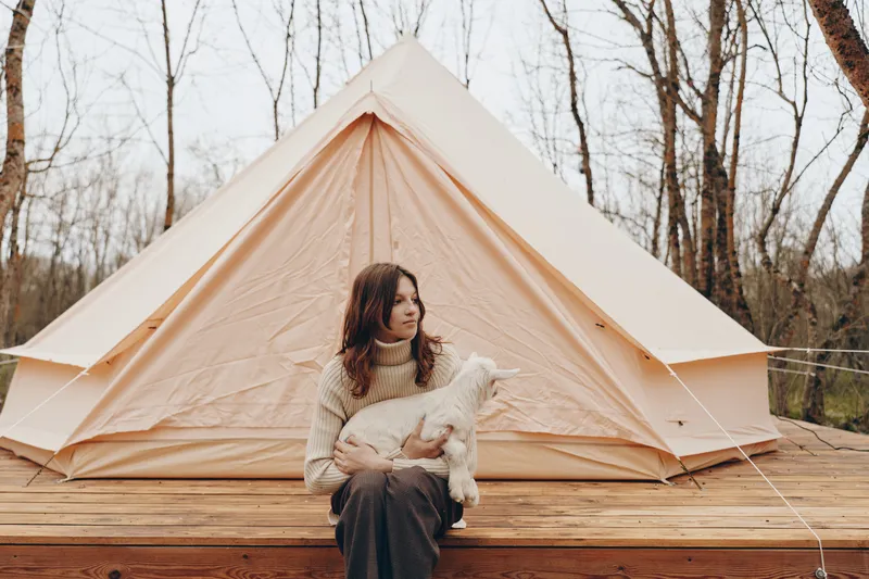 Young woman in a cozy sweater sits on a wooden deck holding a small goat in front of a beige canvas tent in a leafless forest setting during autumn.