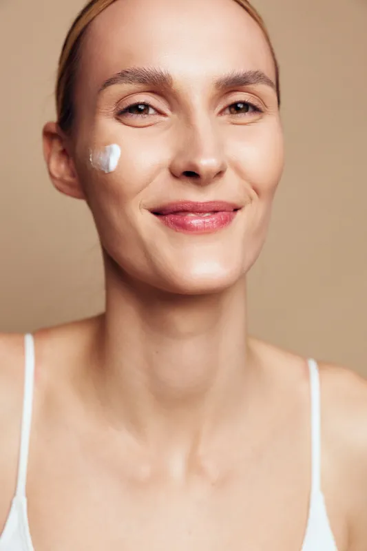 Smiling woman with clear skin applying white face cream on cheek. Close-up portrait on beige background. Concept of skincare, beauty, health, and self-care routine.