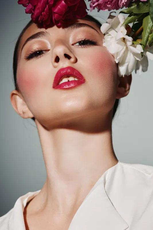 Close-up beauty portrait of woman with glossy red lips and floral headpiece. She wears subtle eye makeup and blush, showcasing fashion and creative cosmetic styling in studio light.