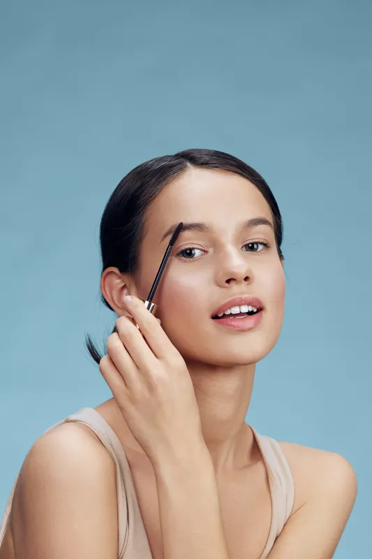 Young woman applying eyebrow gel with a positive expression against a light blue background, highlighting beauty and self care