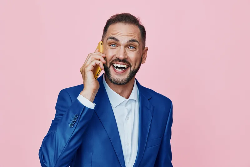A professional man in a blue suit speaks on a mobile phone against a solid pink background, projecting confident energy, friendly business charm, and focused professionalism.