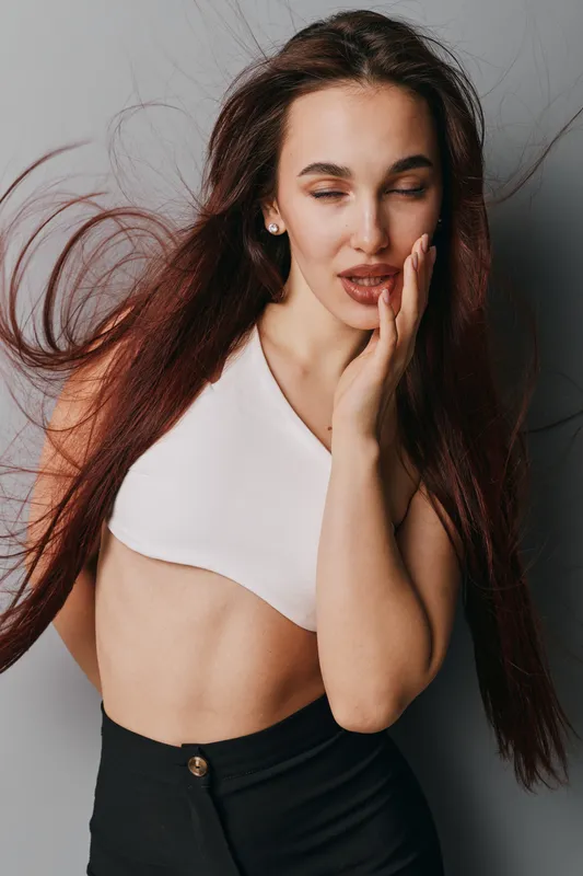 Young woman with long hair posing with closed eyes and hand on face in studio. Fashion model wearing white crop top and black pants with natural makeup and relaxed expression.