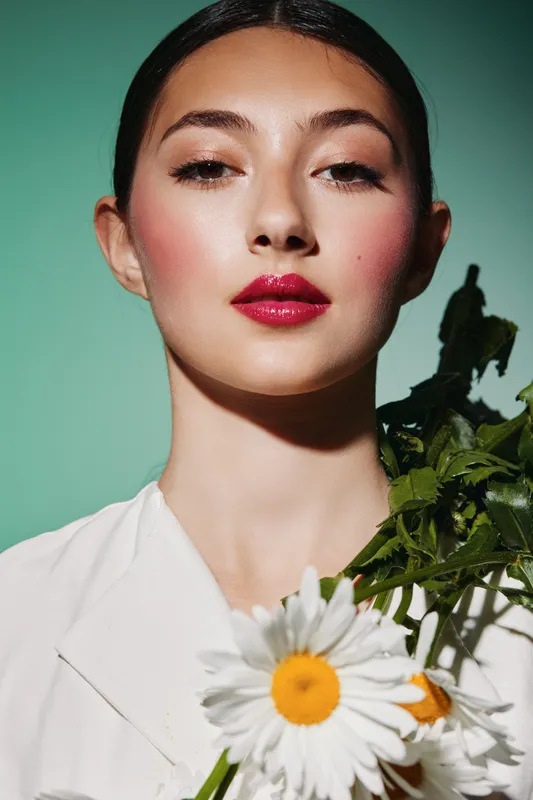 Close-up of confident woman with slicked hair and bold fuchsia lips wearing white jacket holding daisies. Studio fashion portrait on pale green background. Beauty creative concept.