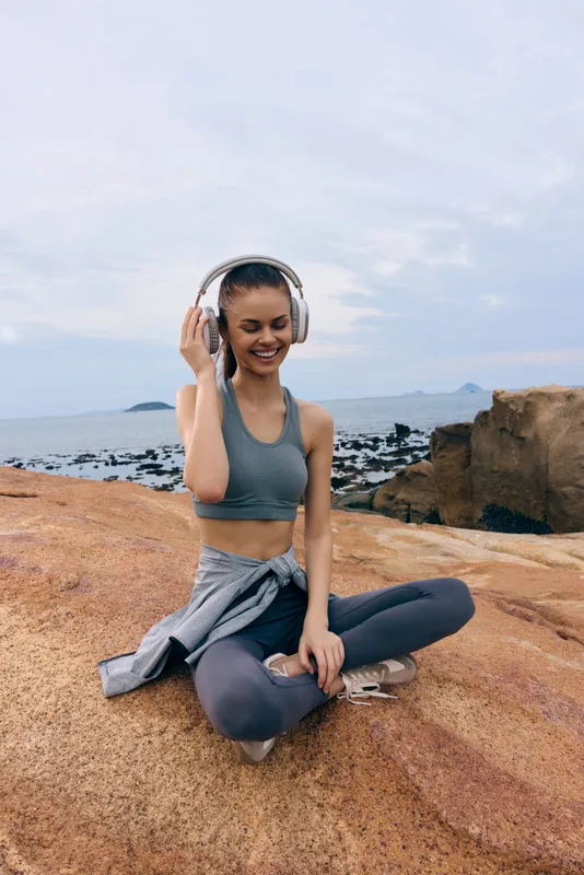 Young woman enjoying music on headphones outdoors, wearing athletic clothes, smiling on rocky beach Ideal for health and fitness concepts