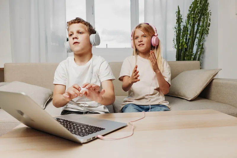Two children with headphones play music on a laptop in a bright living room. The boy uses hand gestures while the girl claps, showing fun and interactive digital entertainment.
