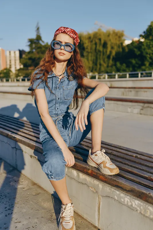 stylish woman in denim jumpsuit and sunglasses sitting outdoors with urban background, casual look, red hair, relaxed pose, sunny day