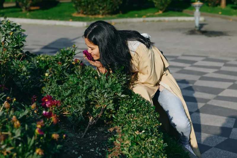 Woman in a light coat leans toward flowering shrubs in a park, smelling flowers and enjoying nature, outdoor gardening and fresh air in a peaceful garden setting, gardening flowers woman outdoor