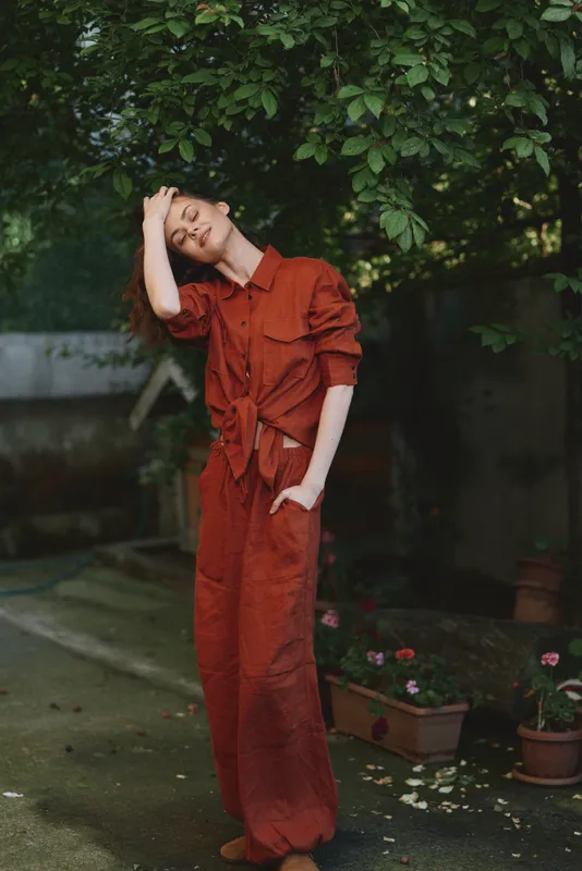 Woman in a red jumpsuit stands in a garden, relaxed pose with hand on head, lifestyle portrait emphasizing casual fashion and outdoor mood